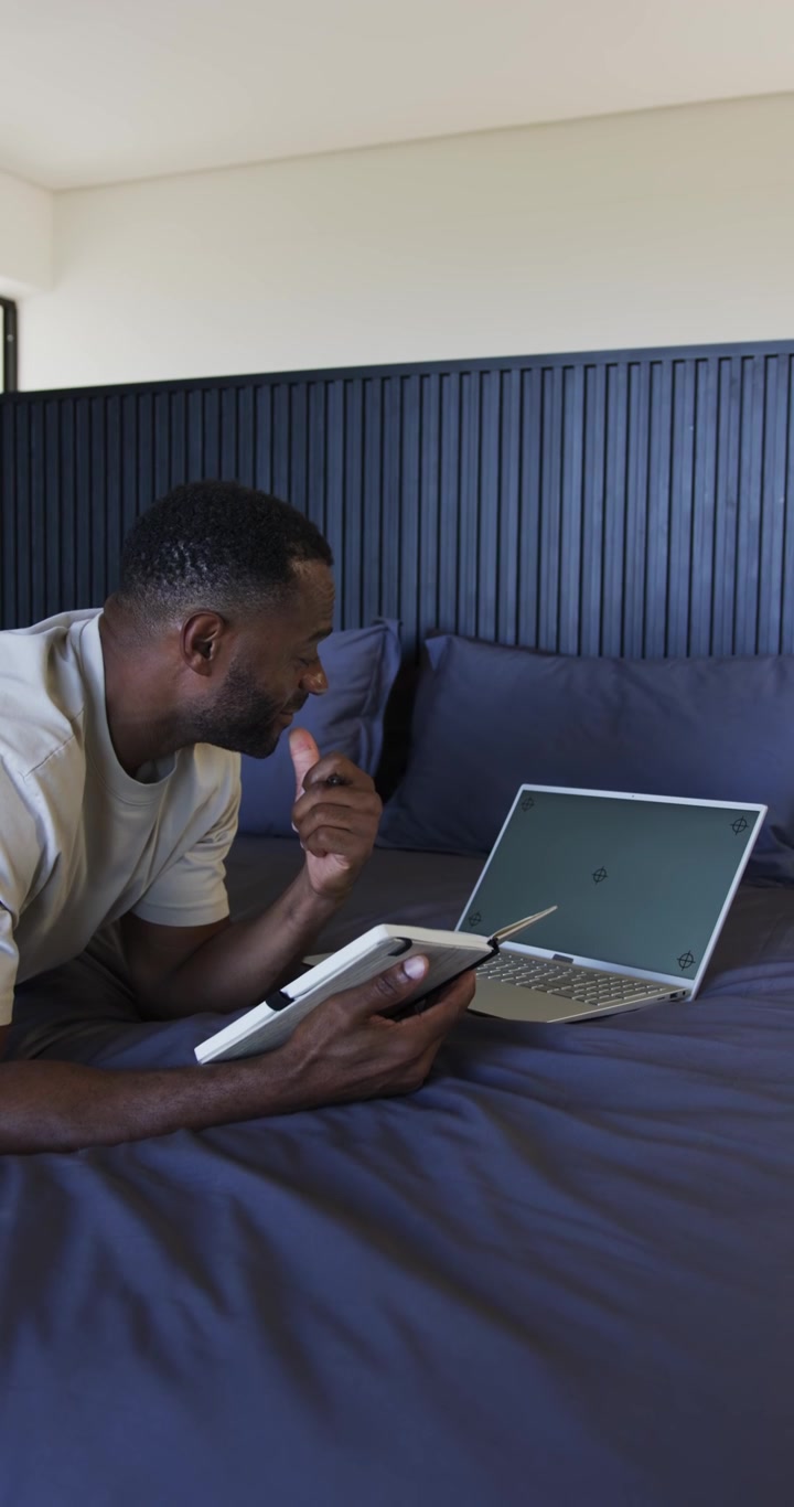 Tapping tablet causing man on bed, holding pink object, glancing to laptop while checking work