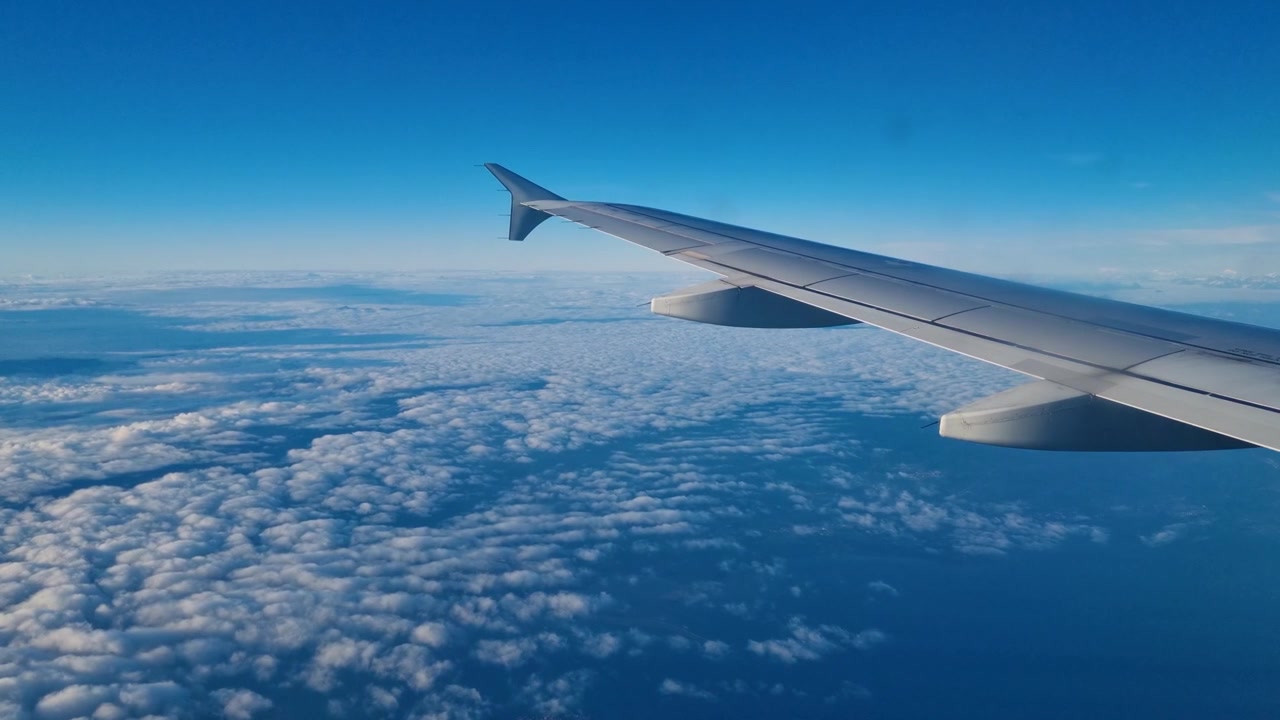 Vista dal finestrino di un aereo che mostra l'ala scivolare dolcemente sopra soffici nuvole bianche sotto un vasto cielo blu, creando un senso di pace, libertà, avventura e viaggio senza fine