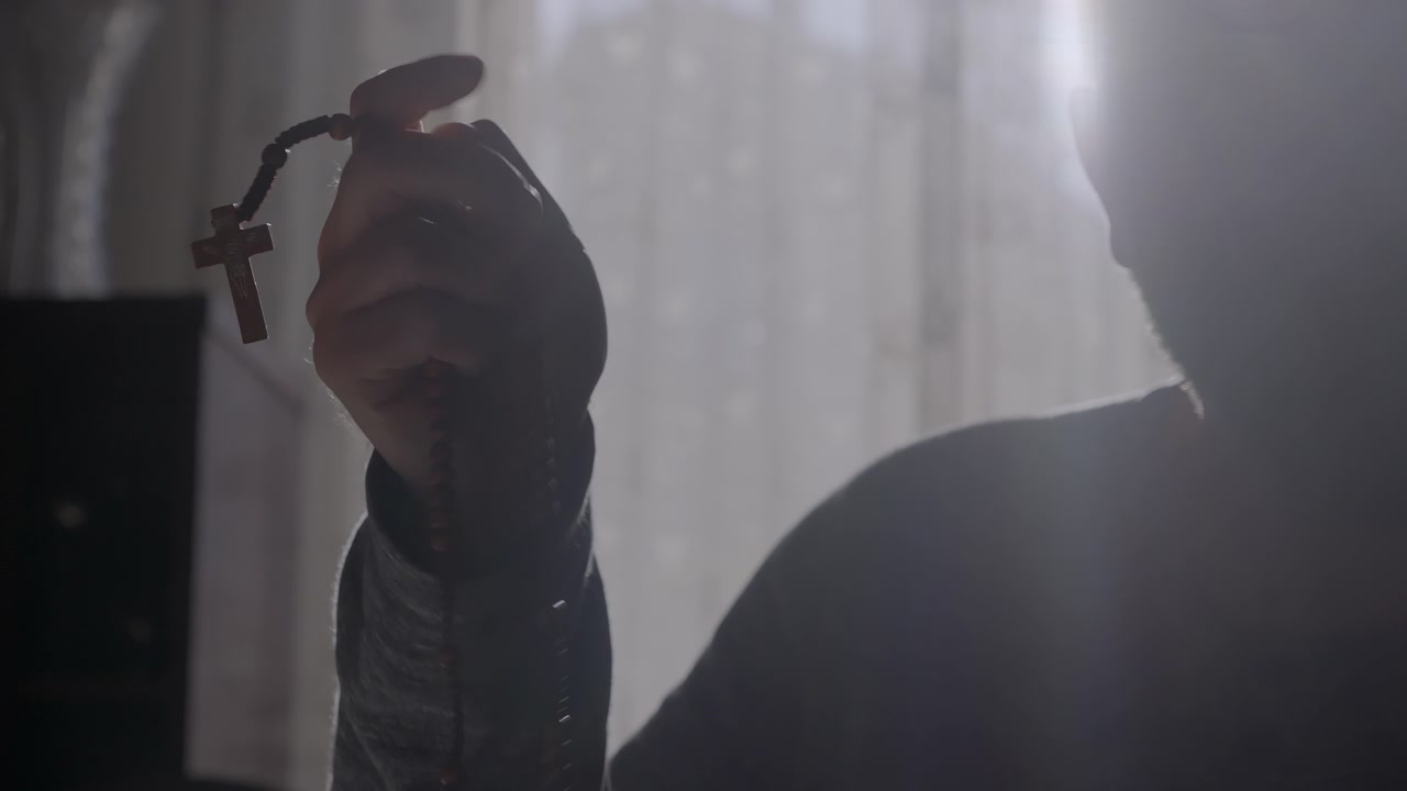 Illuminato da una morbida luce solare, un uomo tiene delicatamente un rosario durante un momento di meditazione. Temi di pace interiore, radicamento spirituale, resilienza e contemplazione