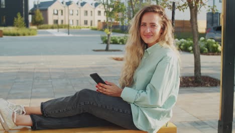 Retrato-De-Una-Mujer-Sonriente-Sentada-En-Un-Columpio-Y-Sosteniendo-Un-Teléfono-Inteligente