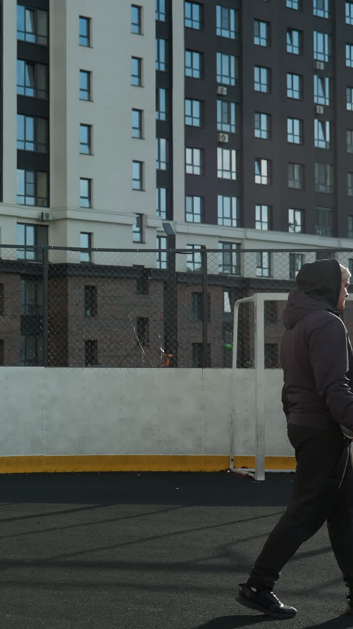 Man in hoodie and beanie cap steps onto football pitch holding ball in right arm with bar net and office buildings in background, showing urban sports environment