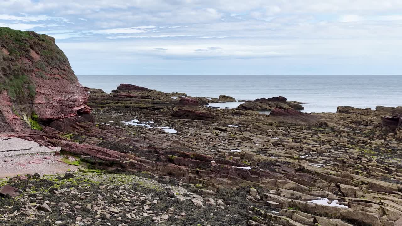 Camera pans over red sandstone cliffs, rocky shoreline, seaweed, and tidal pools at low tide