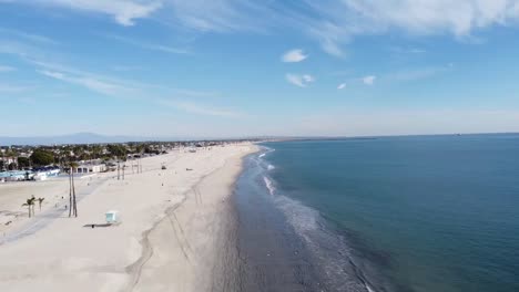 Drone-flies-in-from-ocean-over-beach,-bike-path-and-parking-lot