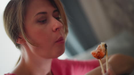 Close up of woman in pink top eating sushi with chopsticks as she savors the flavor in calm setting, soft blur background with curtain and dim light filtering through