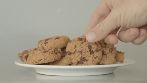 Mano-Agarrando-Galletas-Con-Trocitos-De-Chocolate-En-Un-Plato-Blanco