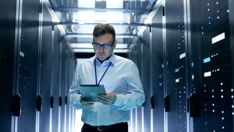 el técnico camina a través de filas de bastidores de servidores en el centro de datos. simultáneamente trabaja en una tableta.