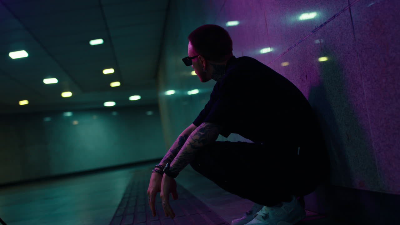 Tattooed man in sunglasses crouching in a neon-lit urban underpass