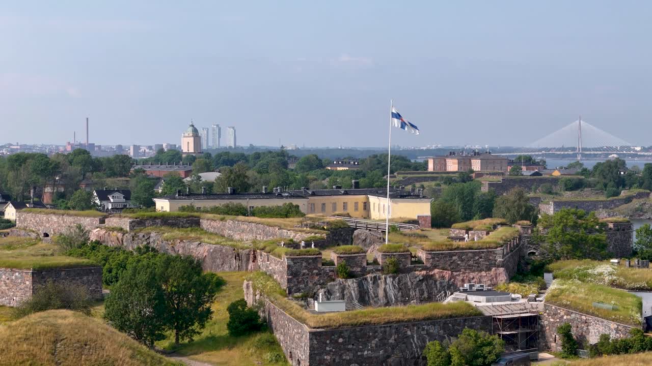 Slow, rising aerial view showing Finnish flag flying above the Suomenlinna sea fortress with city skyline behind it in Helsinki, Finland