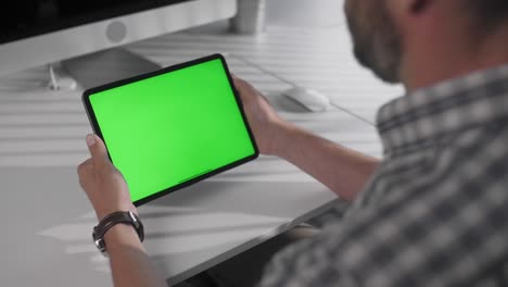 view from the shoulder of man holding and using hand gestures swipe right on green mock-up screen digital tablet computer sitting at the desk. browsing the internet. background office