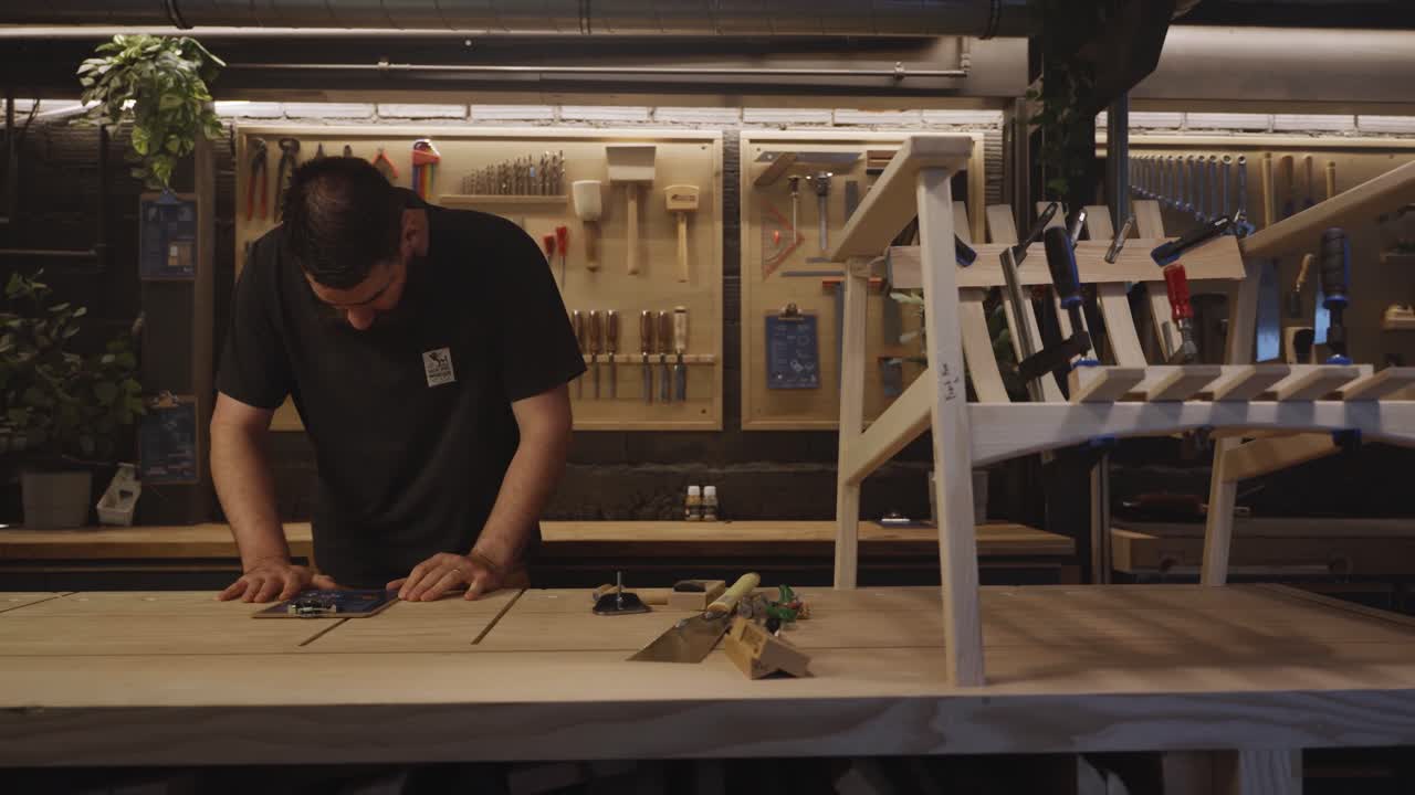 A craftsman stands in a modern woodworking studio surrounded by tools, a workbench, and a wooden furniture frame, representing craftsmanship, creativity, and handmade design