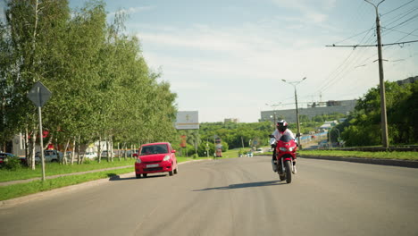 a motorcyclist wearing a helmet rides fast on a highway with a red car seen from behind, with buildings, trees, and distant cars in view