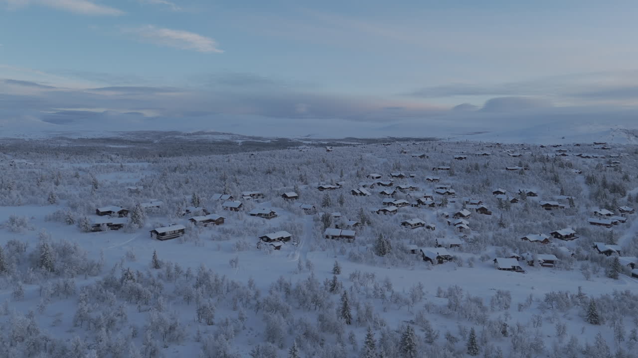 Forwards flying wide droneshot flying over a snowy landscape scene with many small houses in Norway with frozen trees on a bright day during sunrise with clouds in the sky LOG