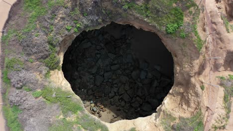 la fascinante toma de un dron captura la vista única de un agujero circular tallado en los acantilados de la playa mientras el dron asciende lentamente, revelando las impresionantes formaciones naturales que lo rodean.