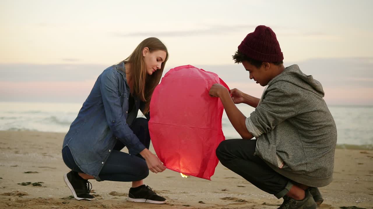 해가 지는 동안 해변에서  ⁇ 만적인 데이트: 젊은 다민족 커플이 발사하기 전에 빨간 종이 등불을 들고 있습니다. 매력적인 여성