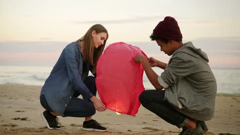 romantic date at the beach during sunset: young multi ethnic couple holding red paper lantern before launching. attractive woman