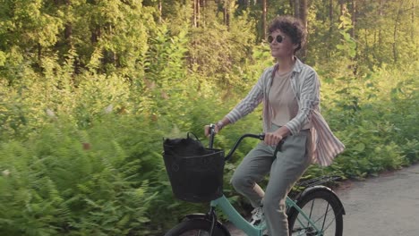 joven feliz con gafas de sol y ropa informal montando en bicicleta en un sendero forestal