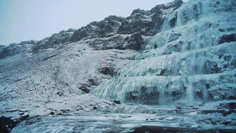 A-beautiful-female-tourist-admiring-the-rocky-waterfall-Dynjandi,-Iceland-on-a-cloudy-day---Wide-pan-shot