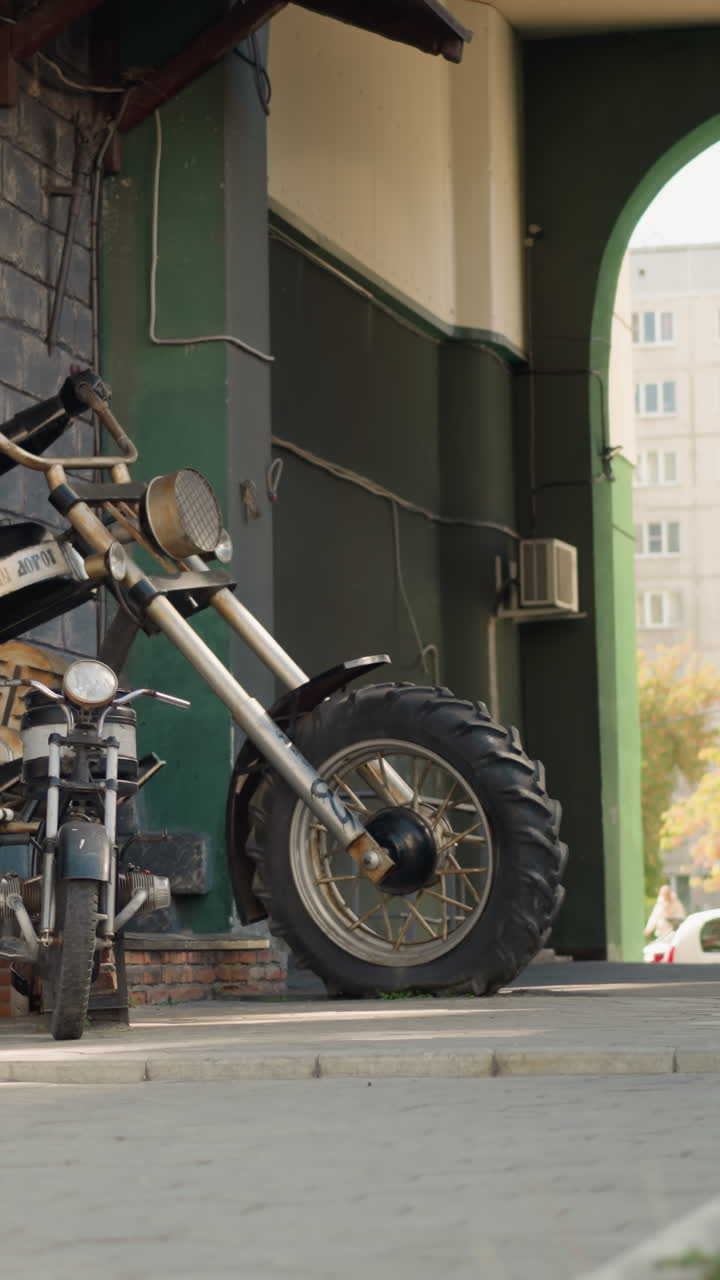 초록색 아치 아래 주차된 헬기 오토바이. 풍화된 커스텀 헬기가 벽돌 벽에 기대어 있고, 부드러운 햇빛을 받는 과도한 크기의 앞바퀴와 크롬 포크, 포장도로 위의 먼지 묻은 타이어, 흩어져 있는 상자들이 보여요.