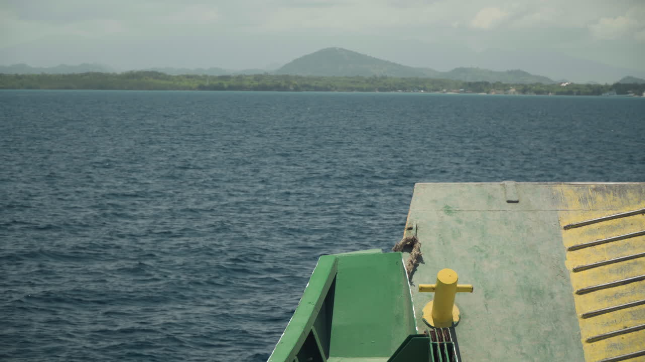 Prua del traghetto con ringhiera verde, bitta gialla oceanica sulla rampa di dritta, orizzonte montuoso con isola distante, cielo coperto, acqua calma, toni tenui, traversata lenta, solitudine nautica, perfetto per i viaggi