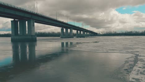 grey bridge over wide river with ice against dense forest