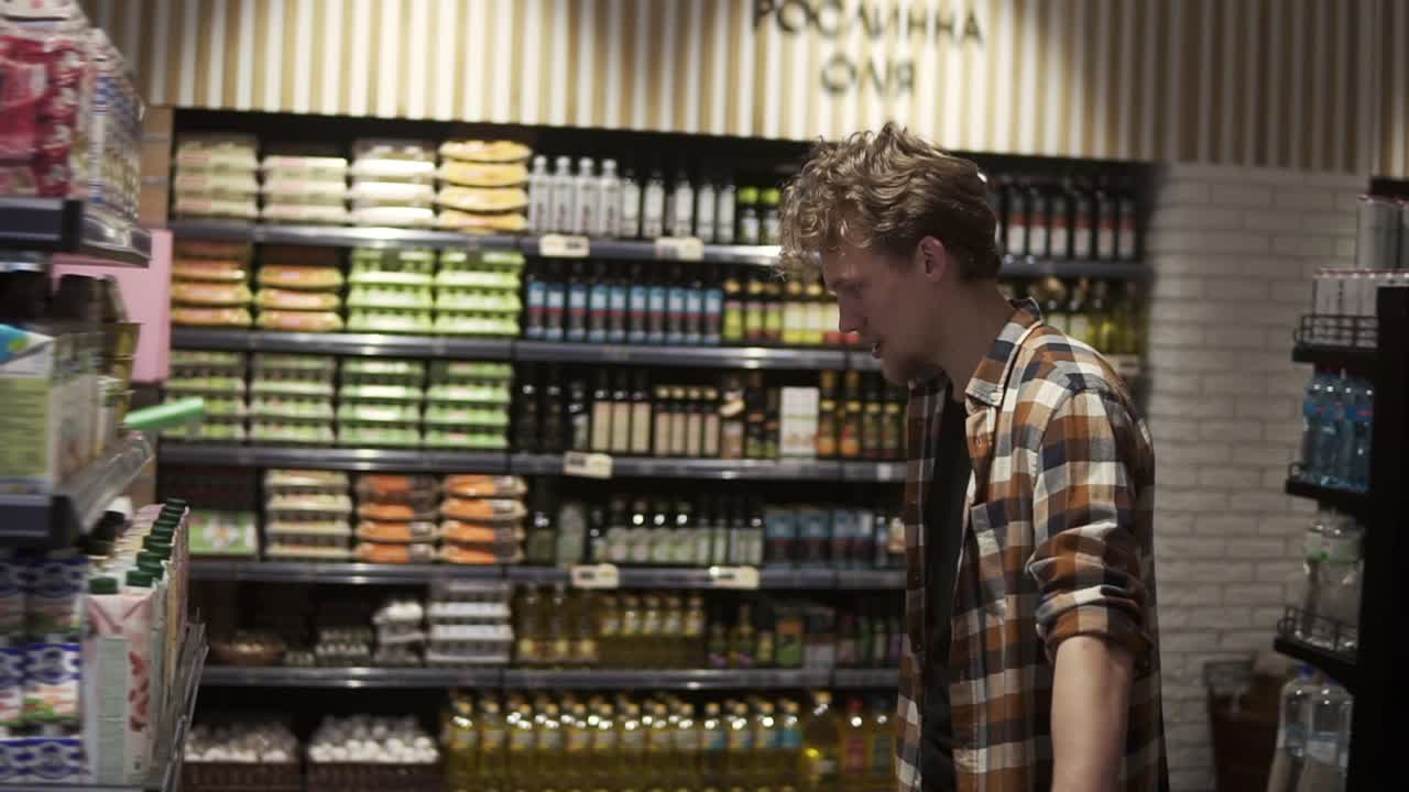 Young guy in supermarket. Caucasian handsome guy in plaid shirt choosing pasteurized milk from variety on the store shelves. Side view