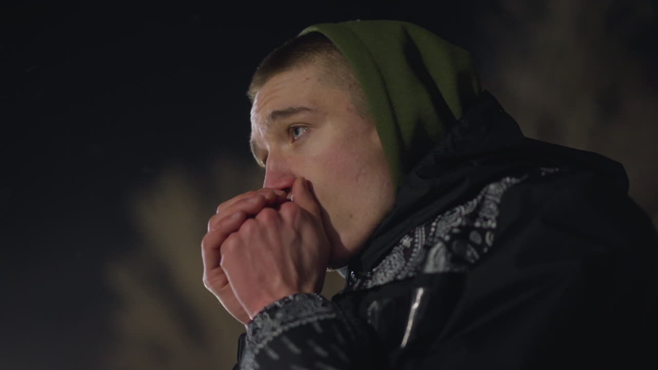 Cold boy in hooded jacket blows warm air into hands to stay warm during freezing night, visible breath rising as blur background and distant light enhance cold atmosphere