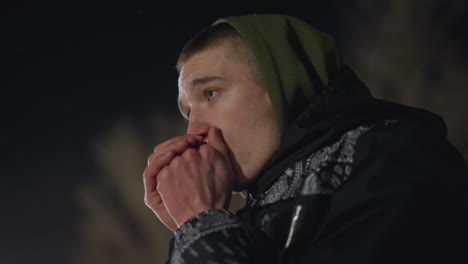 cold boy in hooded jacket blows warm air into hands to stay warm during freezing night, visible breath rising as blur background and distant light enhance cold atmosphere
