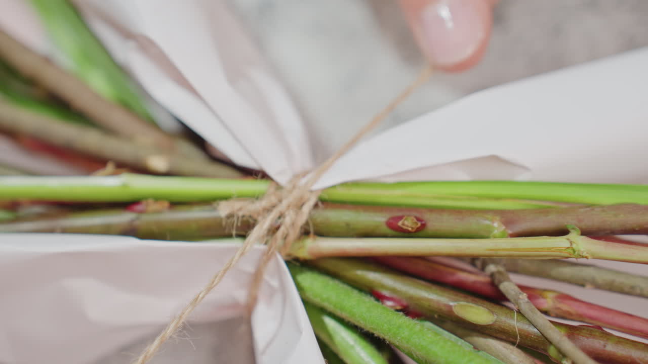 Florist hands tying bouquet stems with twine, closeup showing delicate fingers wrapping paper around green stems, rustic jute knot detail, artisanal workshop atmosphere and careful finishing touch