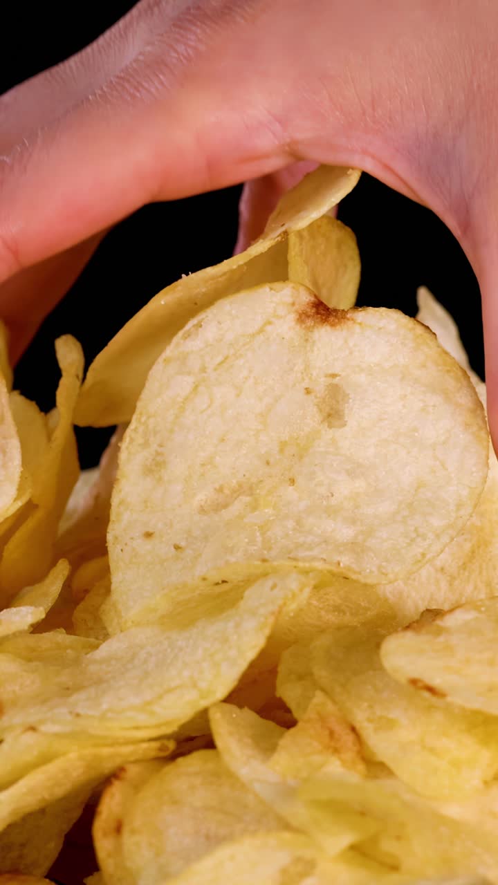 Human hand selects potato chips from pile, close-up, bright lighting, black background, slow camera movement