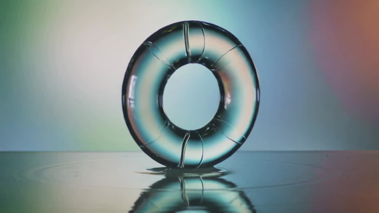 Rotating glass torus tilting above shiny water after thin column touching surface, creating ripples