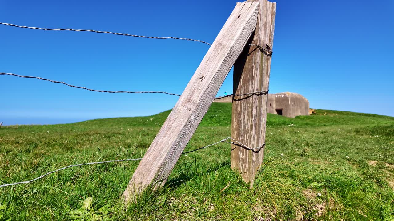 Memorial place with barbed wire fence with wooden support and coastal German bunker in background, Longues-sur-Mer, Normandy, France.