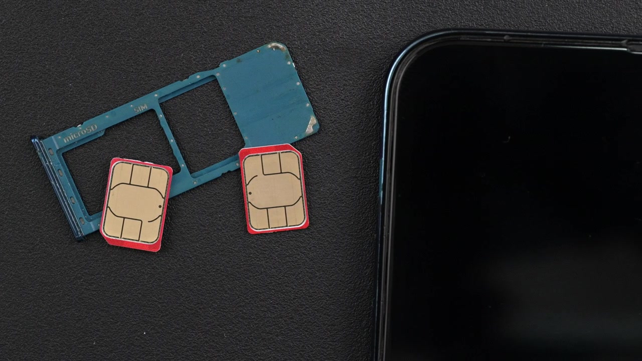 Dois cartões SIM com bordas vermelhas e uma bandeja de cartão SIM azul estão colocados ao lado de um smartphone em uma superfície escura. Útil para análises de tecnologia ou tutoriais de dispositivos móveis