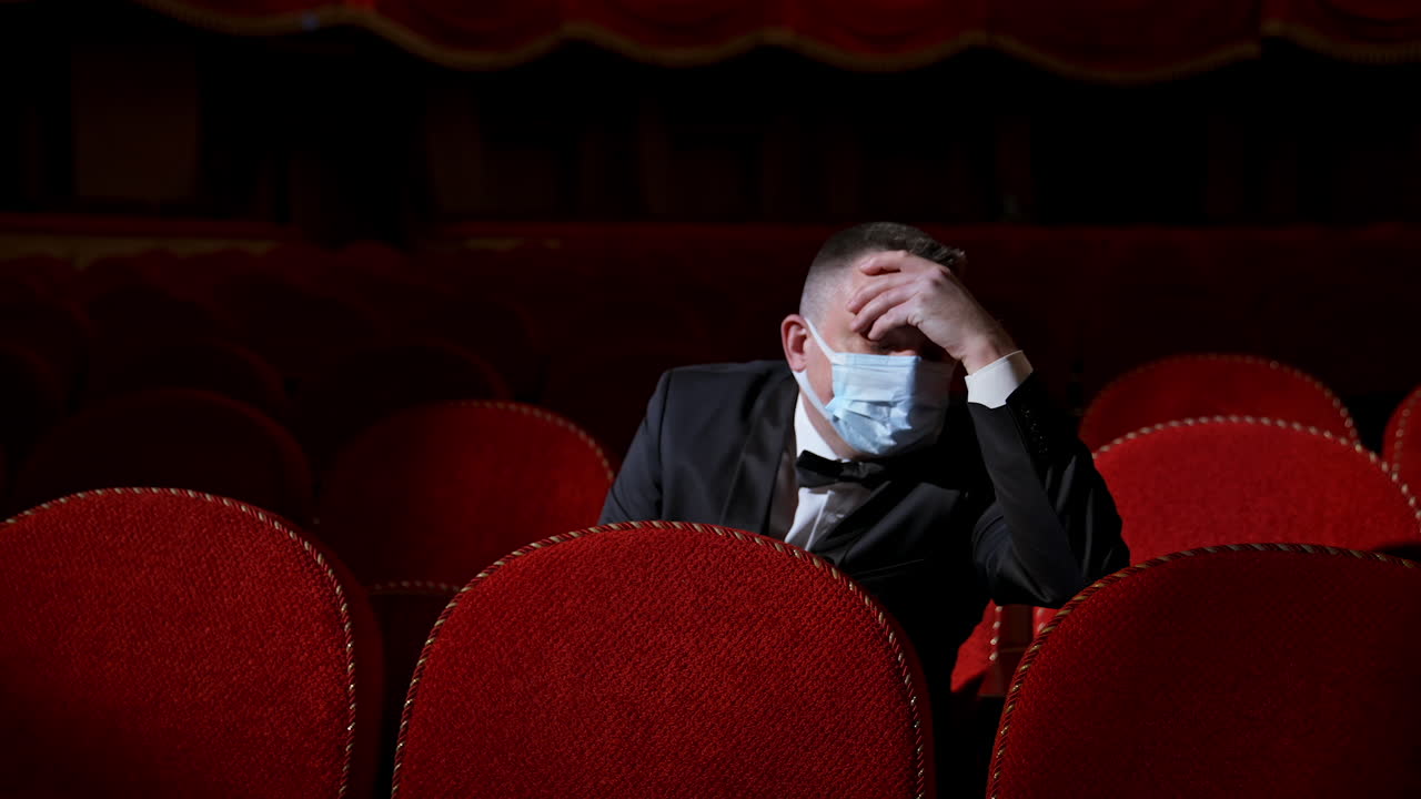Tired businessman in medical mask in empty theater. Man with bad well-being is sitting in auditorium without people. Covid-19.
