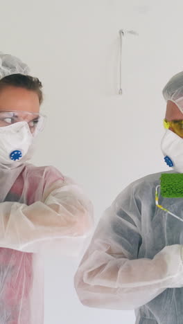 funny young couple wearing protective suits with hoods and masks poses hiding behind rollers in green paint at renewal