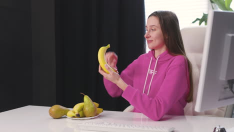 happy young woman sitting at desk peeling and eating banana