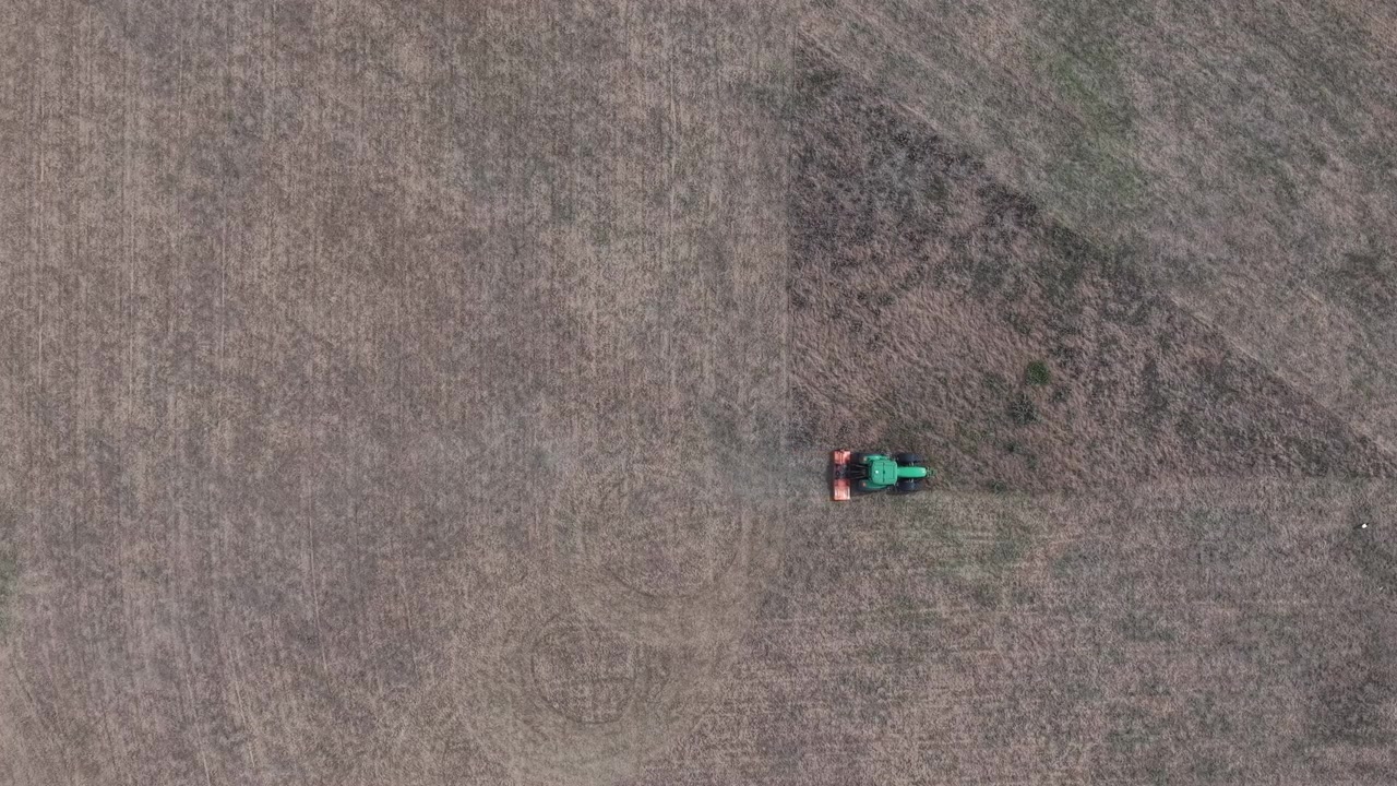 Una vista aerea mostra un trattore che coltiva un grande campo arido, illustrando le moderne pratiche agricole industriali e la preparazione stagionale del terreno.
