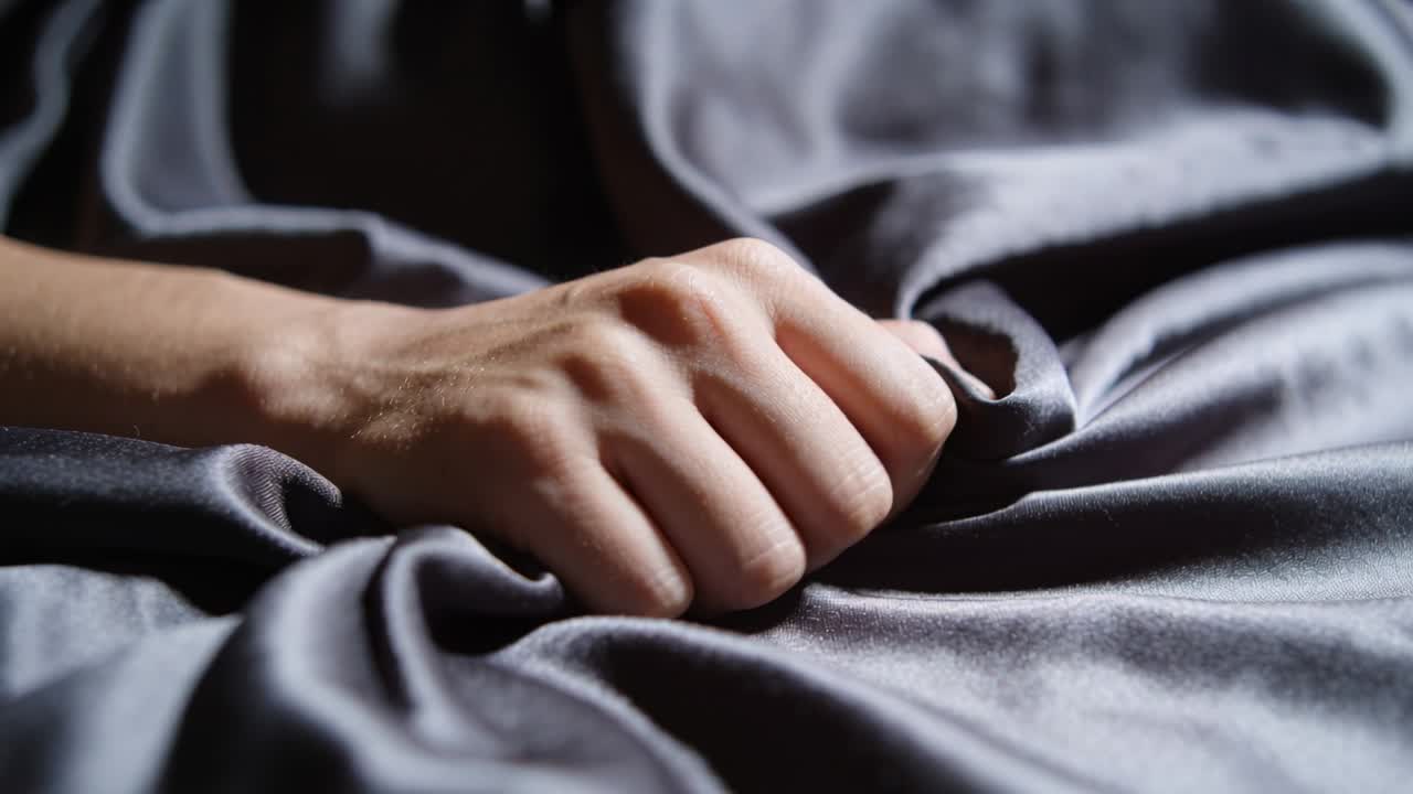Una mano e un avambraccio pizzicano e tirano un lenzuolo di raso scuro sul letto della camera, formando pieghe arricciate radiali.