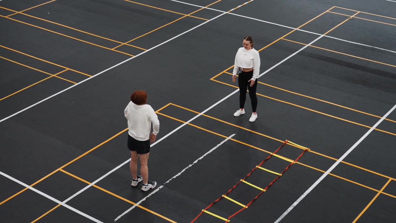 Coach demonstrates shuttle placement and tactics on marked court, instructional dialogue and slow demonstration of angles, attentive trainee watches amid agility ladder and court lines