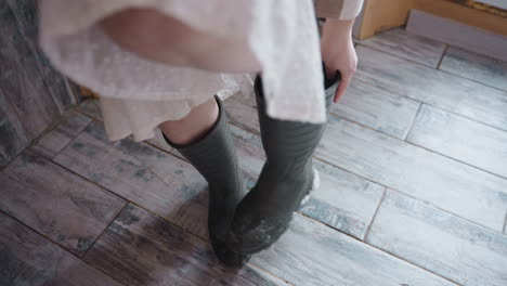 elegant woman removing work boots while standing on rustic tiled floor, ruffled dress hem lifting to reveal bare calves, hands gripping boot edges, wood door frame and frost specks blurred