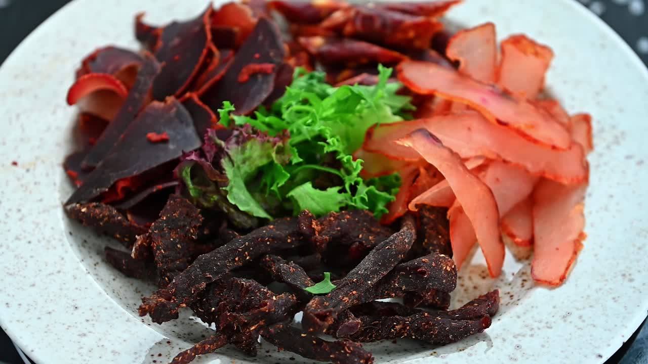 Berbagai jenis daging disajikan di atas piring putih bersama dengan sayuran dan rempah-rempah. Sepiring berisi berbagai daging, termasuk dendeng dan daging sapi kering, dengan sayuran segar yang ditempatkan di sekelilingnya.