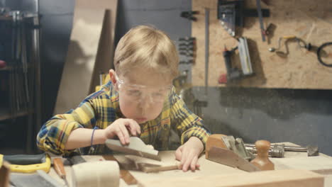 niño pequeño y lindo caucásico con gafas puliendo madera en el taller