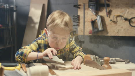 caucasian little cute boy in goggles polishing timber in workshop