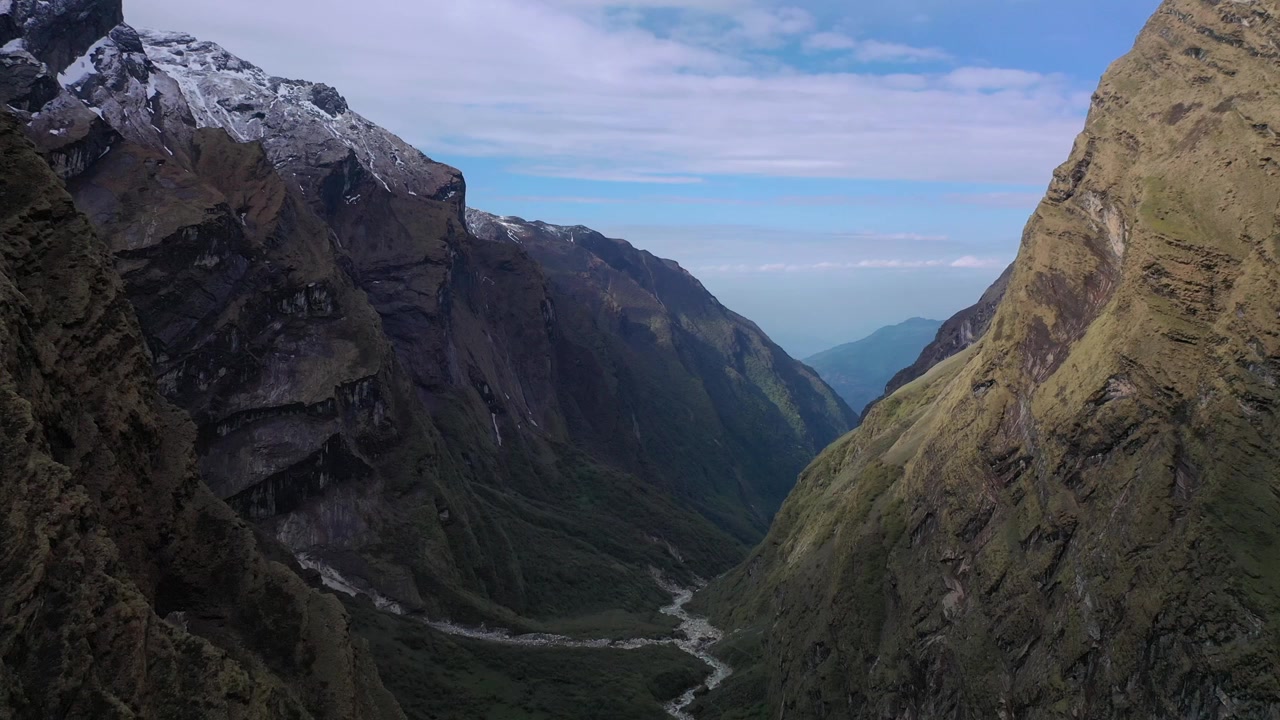 Panoramica mozzafiato di una profonda e aspra gola di montagna in Nepal. Presenta un fiume stretto che serpeggia sul fondovalle, fiancheggiato da imponenti scogliere alpine e ripidi pendii erbosi sotto un cielo azzurro