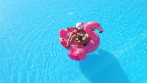 aerial: young tourist couple lies on a pink flamingo floatie during a pool party