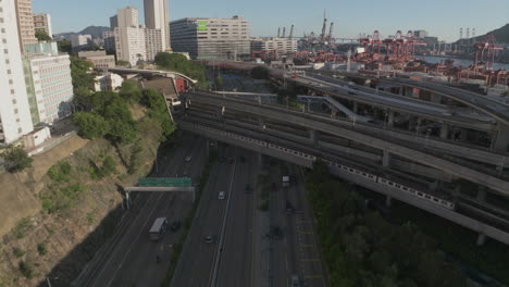 Aerial-forward-flight-over-traffic-on-highway-and-Tsuen-Wan-line-MTR-train-on-bridge-in-Hong-Kong-City---Industrial-Port-in-background-during-sunny-day