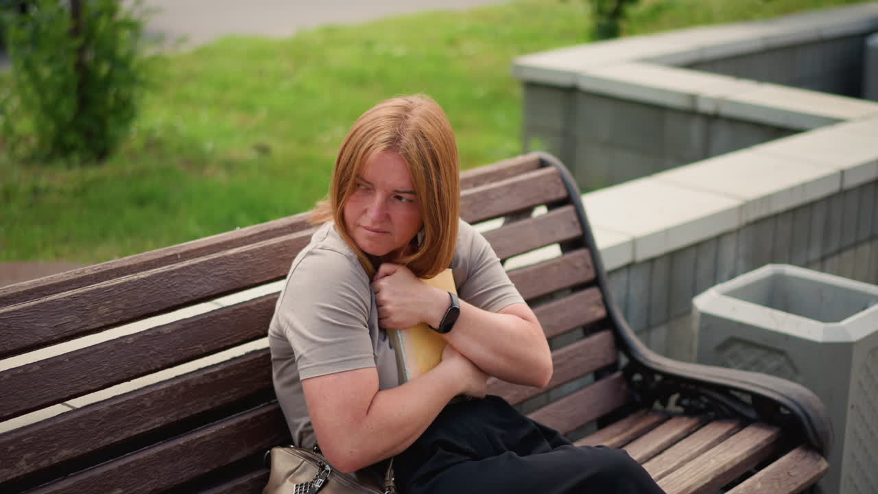 Student sitting on bench outdoors reading book, flipping through pages slowly then closing it and holding against chest with gentle emotion, cars and pigeons in background