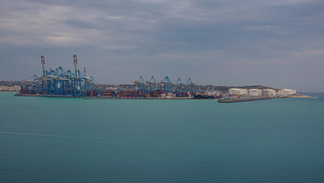 Tiro-De-Lapso-De-Tiempo-De-Una-Gran-Grúa-Azul-Cargando-Un-Gran-Barco-De-Contenedores-En-Kalafrana,-Birzebbuga,-Malta-En-Un-Día-Nublado