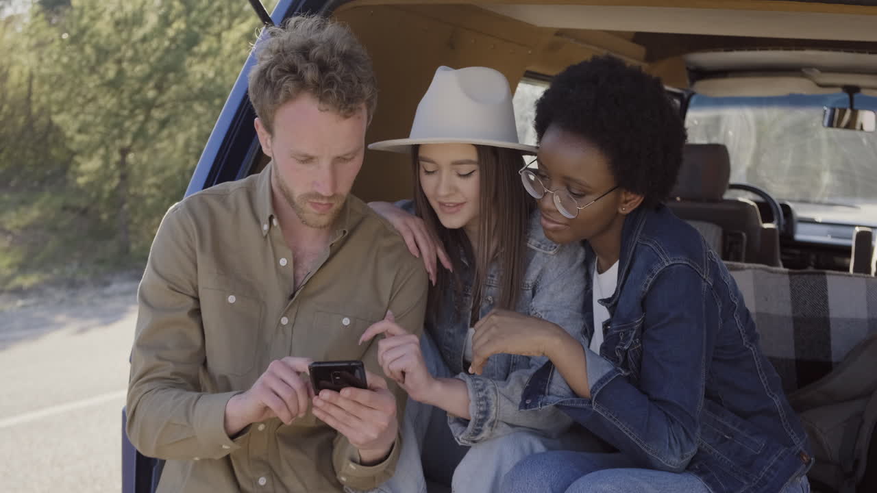 un joven mostrando su teléfono celular a dos hermosas jóvenes dentro de la caravana durante un viaje por carretera 1