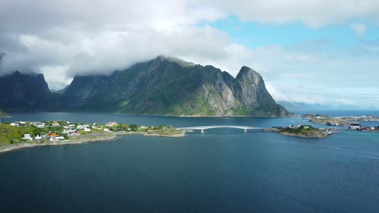 Aerial drone flying over Lofoten large blue lake and towards a small town with houses and a bridge going over water from cliffs. Large mountain in the background with moss and green landscapes.