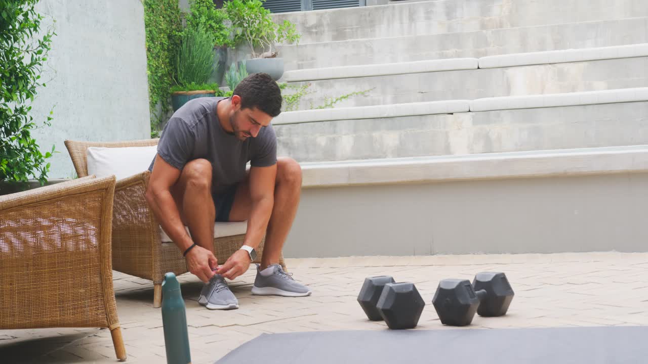 Hombre de veintitantos años atándose y ajustándose los cordones de los zapatos en un banco del patio, preparándose para entrenar con mancuernas.